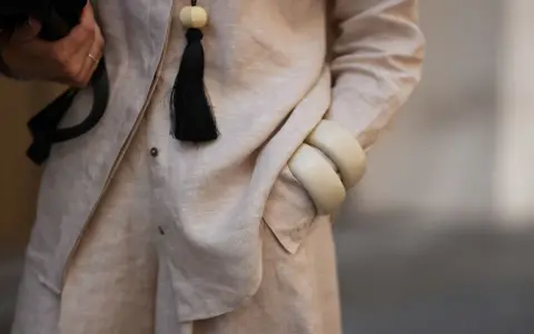 Dettaglio di un outfit in lino materiale, con una mano che tiene una borsa nera, un ciondolo con nappa nera e bracciali color crema.