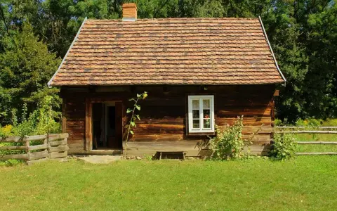 Drewniana chata z glinianym dachem, typowa dla skansenów w Polsce, otoczona zielenią i drewnianym płotem.