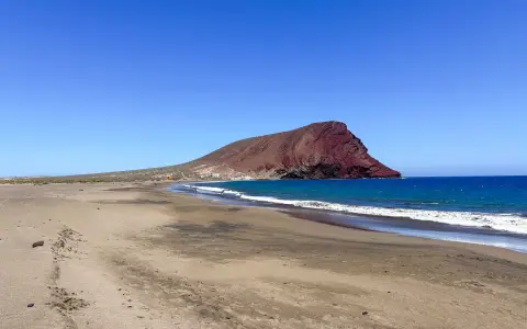Plaże z białym piaskiem na Teneryfie, z charakterystyczną czerwoną górą w tle i turkusowym oceanem.
