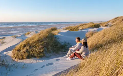 Ett par sitter på en sanddyn och njuter av utsikten över havet. Vackra sanddyner med gräs sträcker sig längs kusten, en idyllisk plats i en nationalpark i Danmark.