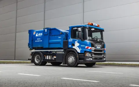 Niebieska śmieciarka Scania z napisem "ODPADY" i logo BYS. Pojazd gotowy do odbioru gruzu, z kodem odpadu na tablicy.