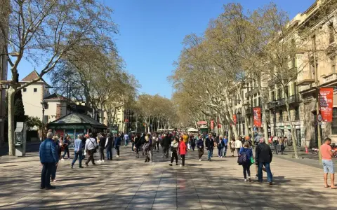 Tłum ludzi spaceruje po słonecznej La Rambla w Barcelonie, otoczonej drzewami i zabytkowymi budynkami.