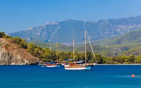 Dwa drewniane jachty kołyszą się na turkusowych wodach, zalesione wzgórza i góry w tle. Idealny dzień na zwiedzanie wybrzeża w okolicach **Antalya miasto**.