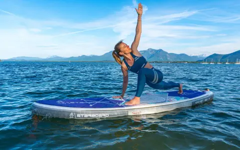 Frau praktiziert Krieger II auf einem SUP-Board auf ruhigem Wasser, Berge im Hintergrund.