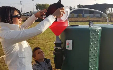 Une femme verse des déchets organiques dans un entonnoir rouge, alimentant un méthaniseur domestique. Un garçon regarde, curieux.
