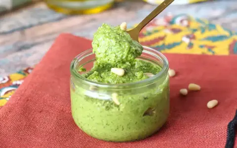 Łyżka nabiera aromatyczne pesto z rukoli z małego słoiczka. Na czerwonym tle widać orzeszki piniowe.
