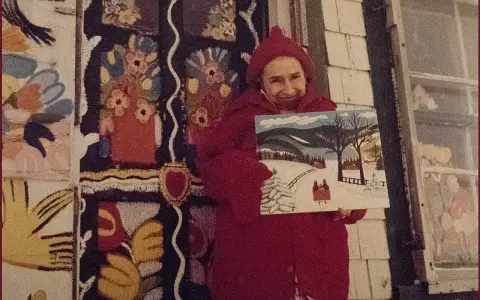 Maud Lewis, klädd i rött, håller upp en av sina charmiga vintermålningar framför sitt färgstarka hem.