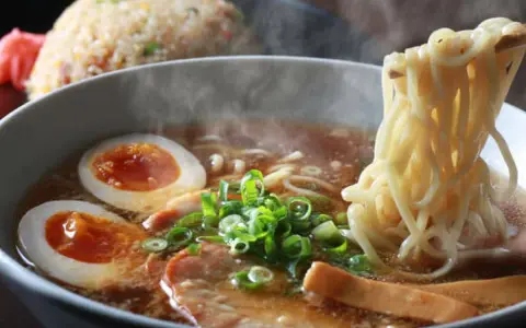Un humeante tazón de ramen con huevo cocido, cebolleta y cerdo. Los palillos levantan fideos, listos para disfrutar de esta deliciosa receta ramen.