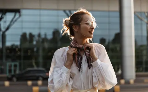 Uśmiechnięta kobieta w okularach poprawia apaszkę na szyi. Pokazuje, jak zawiązać apaszkę, tworząc stylowy look.