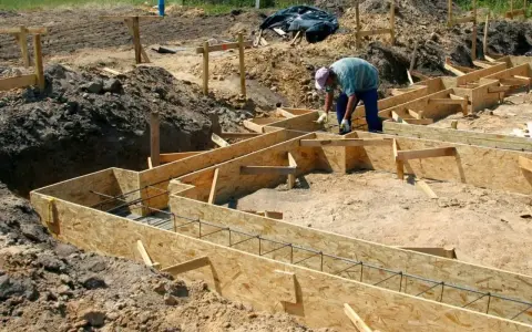 Budowa fundamentów: pracownik układa zbrojenie w drewnianym szalunku. Widać wiele desek na szalunek fundamentu, gotowych do zalania betonem.