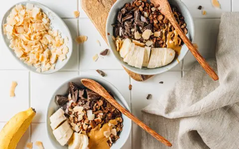 Due ciotole con granola fatta in casa, banana a fette, cioccolato fondente e burro d'arachidi. Cocco in scaglie a lato.