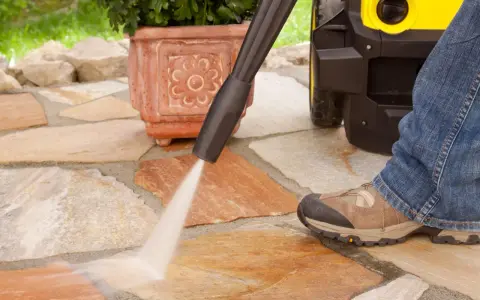 Pulire pavimento esterno con idropulitrice. Un getto d'acqua potente rimuove lo sporco dalle pietre del patio, mentre una persona in jeans e scarponi osserva.