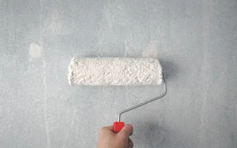 Main tenant, comment rattraper une peinture mal faite. Un rouleau à peinture blanc est tenu par une main sur un mur bleu clair texturé.
