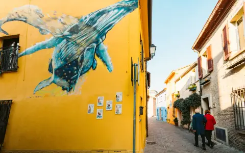 Un murale di balene blu su un muro giallo a San Giuliano Rimini. Una coppia passeggia lungo la strada acciottolata.