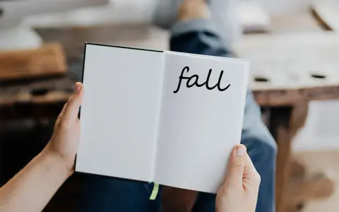 Otwarta książka z napisem "fall" na prawej stronie. Osoba w dżinsach trzyma ją, przygotowując się do jesiennych zapisków.