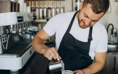 Uśmiechnięty barista w czarnym fartuchu nalewa mleko do kawy. Obok stoi profesjonalna ekspres do kawy.