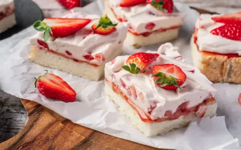 Saftige Stücke eines Erdbeerkuchen Blechs mit frischen Erdbeeren und Sahne, auf Backpapier angerichtet.
