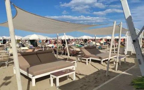 Un lido a Riccione con lettini doppi e ombrelloni bianchi sotto un cielo azzurro e nuvoloso.
