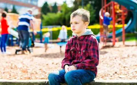Chłopiec z zespołem Aspergera siedzi samotnie na placu zabaw, podczas gdy inne dzieci się bawią.