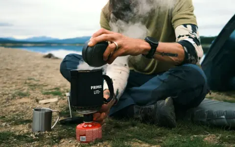 Varm dryck från en gasollykta vid en sjö. En person sitter vid en tältplats och förbereder en kopp.