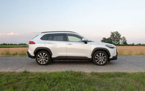 Biała Toyota Corolla Cross Hybrid stoi na poboczu drogi, otoczona polami i zieloną trawą.