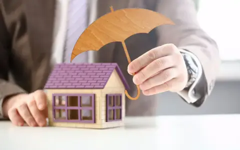 Main d'un homme tenant un parapluie en bois au-dessus d'une maison miniature. Une image symbolisant l'assurance habitation obligatoire.