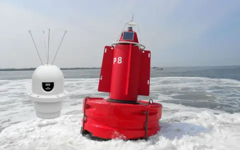 Deux bouées, une blanche avec des antennes et une rouge marquée "P8", flottent sur l'eau agitée, un petit bateau à l'horizon.