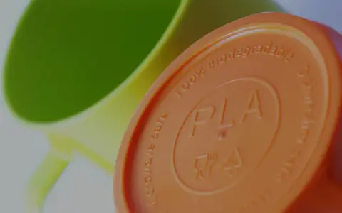 Pomarańczowa pokrywka z napisem PLA, 100% biodegradowalna, obok zielonego kubka.