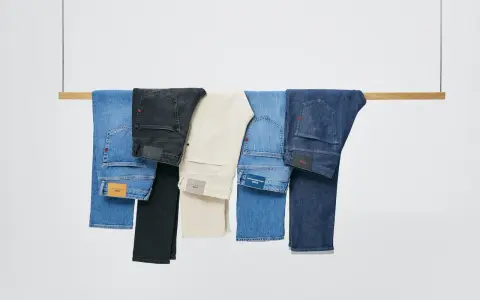 Cinco pares de pantalones vaqueros de distintos tonos cuelgan de una barra de madera. El **denim material** se presenta en azul claro, negro, beige, azul medio y azul oscuro.