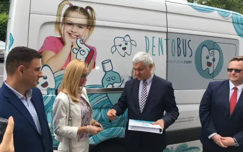 Ludzie stojący przed białym Dentobusem z grafikami uśmiechniętych zębów i dziewczynki.