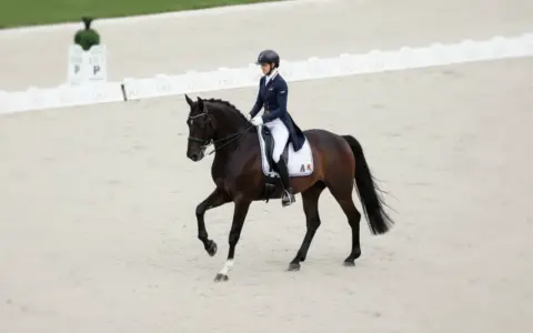 Une cavalière en tenue d'équitation exécute une figure sur son cheval brun. Elle fait partie de la liste des cavaliers français en compétition.