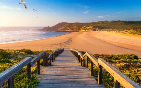 Holzsteg führt zu einem goldenen Strand in Aljezur, Portugal. Möwen kreisen über dem Meer unter blauem Himmel.