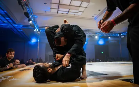 Dwóch mężczyzn w czarnych kimonach walczy w parterze. Intensywna walka jiu jitsu, z widownią w tle.