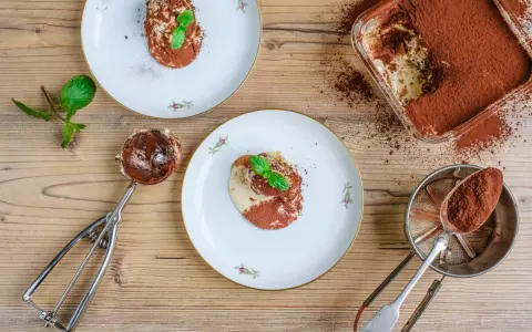 Tiramisu anrichten: Ein Löffel voll cremigen Tiramisus, bestäubt mit Kakao, wartet auf seinen Einsatz. Daneben Teller mit dem fertigen Dessert, garniert mit Minze.