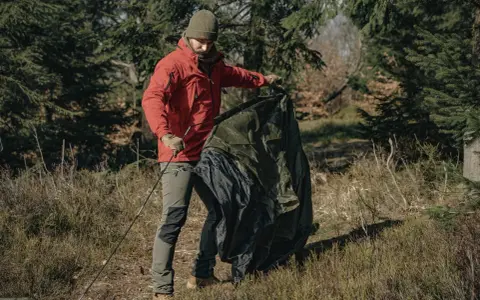 Mężczyzna w czerwonej kurtce składa zielony namiot w lesie. Po biwaku czas na **pranie namiotu**.