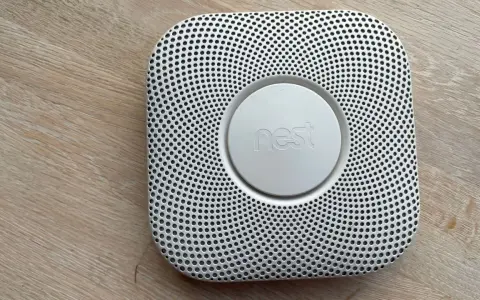 En vit Google Nest Protect-rökdetektor med ett runt "nest"-märke i mitten, placerad på ett trägolv.