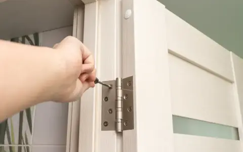 Dłoń dokręca śrubę zawiasu, montując drzwi ukryte w ścianie.
