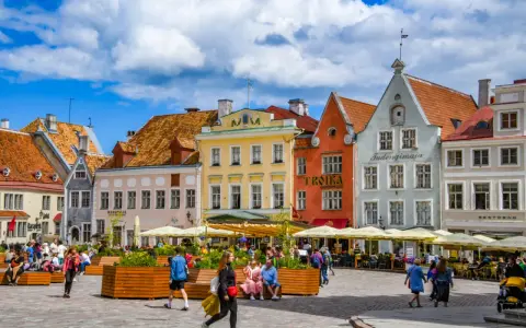 Kolorowe kamienice na placu w Tallinie, stare miasto tętni życiem. Ludzie spacerują, siedzą w kawiarniach pod żółtymi parasolami.