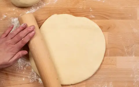 Hand rollt Teig auf Holzbrett aus. Vielleicht mit Dinkelmehl? Was ist Dinkelmehl?