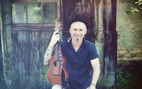 Uśmiechnięty mężczyzna w kapeluszu trzyma ukulele, gotowy do grania nim zajdzie słońce.