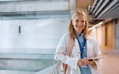 Uśmiechnięta studentka medycyny w białym kitlu, z plecakiem i stetoskopem, trzyma tablet. Zastanawia się, gdzie najłatwiej dostać się na medycynę.