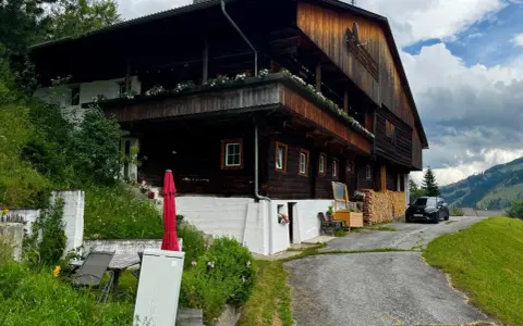 Drewniana austeria w górach, z balkonami ozdobionymi kwiatami. Obok stoi czarny samochód, a w oddali widać zalesione wzgórza.