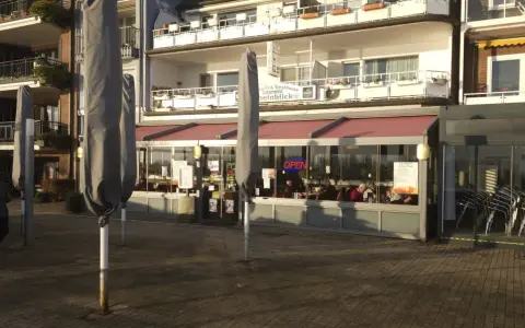 Geschlossene Sonnenschirme säumen die rheinpromenade emmerich. Ein Restaurant mit "OPEN"-Schild lädt zum Verweilen ein.