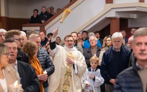 Kapłan błogosławi wiernych podczas liturgii Wielkiej Soboty, unosząc kropidło. W tłumie widać ludzi z zapalonymi świecami.