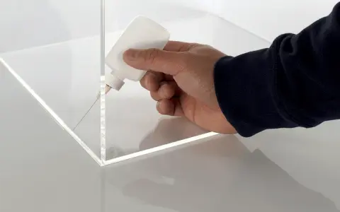 A hand applies adhesive to the corner of a clear plastic box, demonstrating precise gluing of plastic for a clean finish.