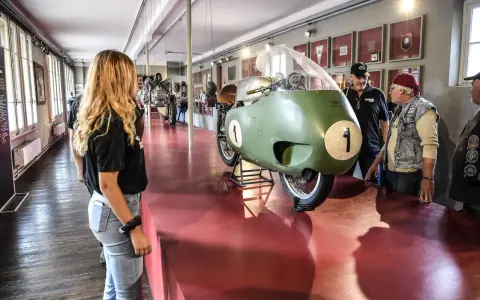 Un raduno moto Guzzi in un museo. Una moto da corsa verde con il numero 1 in primo piano, ammirata da visitatori.