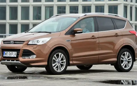 Brązowy Ford Kuga 2019 z kierowcą w okularach, zaparkowany na mokrym parkingu przed nowoczesnym budynkiem.