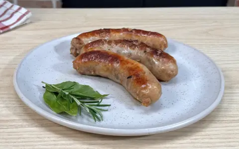 Tre salsicce dorate e succulente, appena cotte in padella, riposano su un piatto con rametti di rosmarino e alloro.