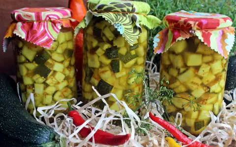 Słoiki z cukinią w zalewie słodko-kwaśnej z curry, ozdobione kolorowymi materiałami. Obok leżą świeże cukinie i papryczki chili.