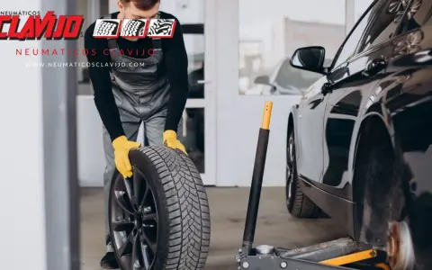 Mecánico con guantes amarillos sosteniendo una rueda, listo para cambiar neumáticos. El coche está en un taller.